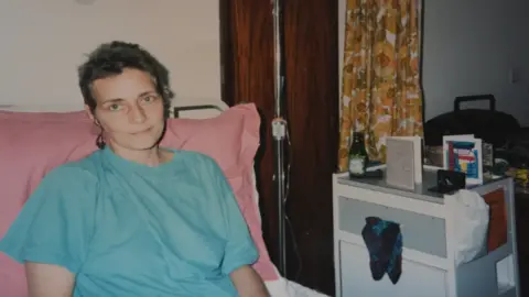 Carolyn Challis laid on a hospital bed during cancer treatment. She is wearing a blue t-shirt and appears to be hooked up to a medical drip. She is looking at the camera.