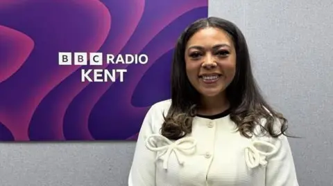 An image of Tia Davies stood in front of the BBC Radio Kent logo. Tia is wearing a white jacket with bows. 