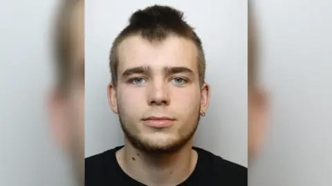 Louis Tavener head and shoulders photo taken after his arrest. He is wearing a black t-shirt, has brown hair, blue eyes and an earing in his left ear.