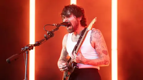 PA Media Simon Neil of Biffy Clyro onstage -he is wearing a vest while playing the guitar and singing into a microphone.
He is bearded and has tattoos over his arms.