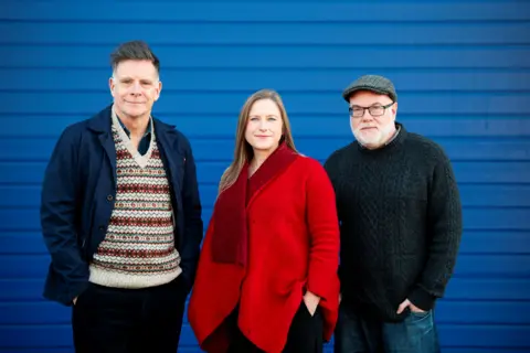 Tommy Ga-Ken Wan Ricky Ross, left, wearing a multi-coloured top under a dark blue jacket. Frances Poet, centre, wearing a red top. Dominic Hill, right, wearing a multi-coloured dark flat cap, glasses and a dark jumper. All three are standing in front of a blue background.