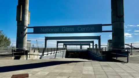 BBC National Glass Centre in Sunderland. Two concrete pillars stand on either side of the centre's wide entrance, which runs into the ground. A sign with the centre's name hangs between the two pillars.