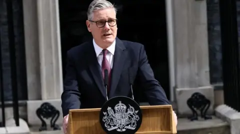 Sir Keir Starmer speaks at a wooden podium in Downing Street. He is wear a dark-coloured suit, white shirt, patterned tie and glasses.