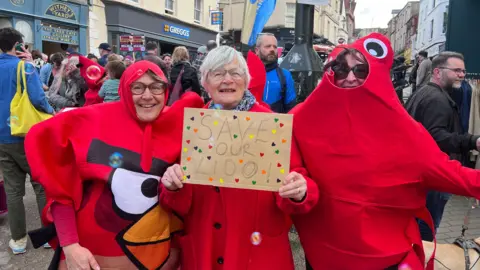 Three women in red fancy dress, pretending to be lobsters, smiling at the camera. One woman in the centre has grey hair and glasses and is holding up a sign which reads 'Save our Lido!'