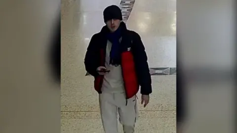 A CCTV image of a man in a grey tracksuit, black and red coat, dark scarf and black beanie. He is looking up and holding a phone.