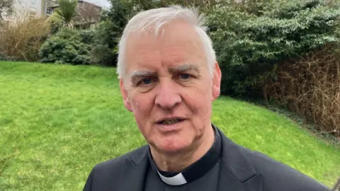 A priest in clerical clothing standing in a garden
