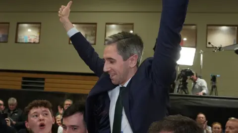 Simon Harris in blue suit jacket, raising arms in celebration as he is reelected, crowd holding him in the air