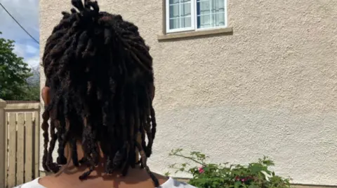 Shows the back of a woman with her hair in dreadlocks and wearing a white t-shirt looking towards a gable wall