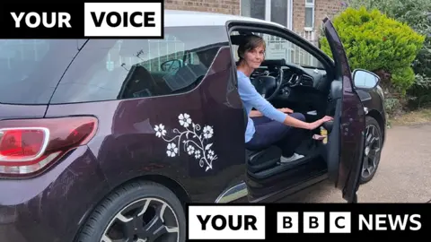BBC Lisa Shackleton sits in the driving seat of her purple Citroen DS3 car with the door open, looking back at the camera, the car is parked on a driveway with bushes in the background, and the words 'your voice, your BBC News' overlaid as a black and white graphic on the image.