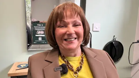A woman with short hair sat in front of a mirror smiling into the camera