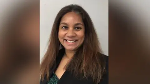 A woman with shoulder-length brown hair smiles into the camera. She is wearing a black blazer and green top.