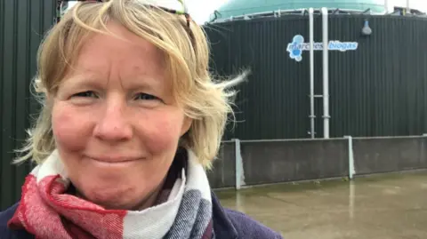 There is a woman with blonde short hair. She is wearing a red, white and navy scarf and has glasses on her head. She is stood in front of a green silo with 'marches biogas' written on the side.