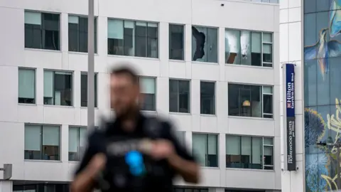 An image showing a building with two broken windows shattered by gunfire. In the forefront is a blurred outline of a Montreal police officer. 