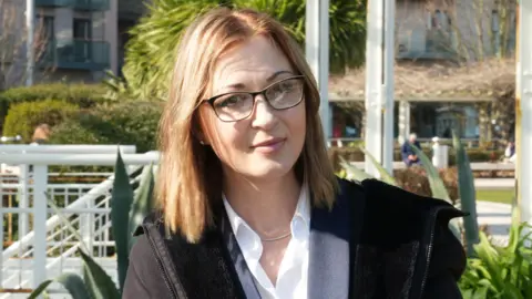 Natalia Baker is looking at the camera with her head slightly angled. She is sitting outside in a park that has pathways and planting. She has shoulder-length blonde and brown hair and is wearing glasses. She is wearing a black coat over a navy jacket with a white shirt underneath. 