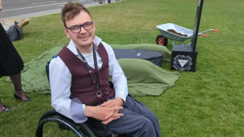 George sits in his wheelchair in front of a mock grave