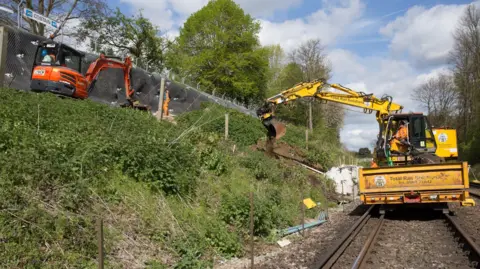 Network Rail A small yellow crane on a railway line working on an embankment, which has a small orange crane on.