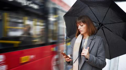 A woman holding an umbrella checks her phone as a bus drives past