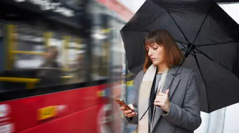 Getty Images A woman holding an umbrella checks her phone as a bus drives past