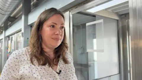 A woman with brown hair is wearing a white shirt. She is standing next to a glass wall.