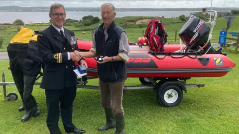 Fleet Bay Inshore Rescue Service Original crew member, Kerr McConchie wearing wellies, brown trousers and a blue gilet receiving the Queens medal from a man wearing a black military uniform , standing in front of the current red lifeboat 