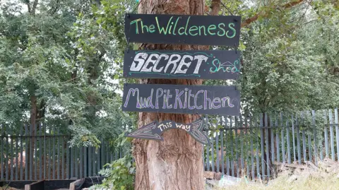 Gatis Community Space A tree with four handwritten signs on it. The rectangular blackboards say 'The Wilderness', 'Secret Shed', 'MudPit Kitchen' and the four sign undernearh says 'This Way' with an arrow pointing to the left. Behind the tree is a grey metal fence and trees. 