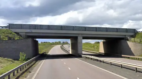 A concrete bridge with a dual carriageway underneath. There are grass banks on each side of the road. 