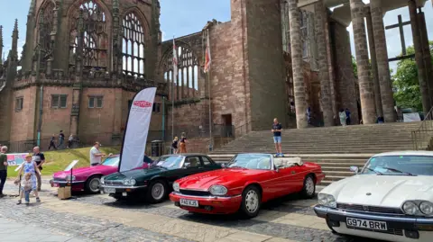 Four XJS cars, coloured white, red, black and pink, are parked outside Coventry's cathedral ruins