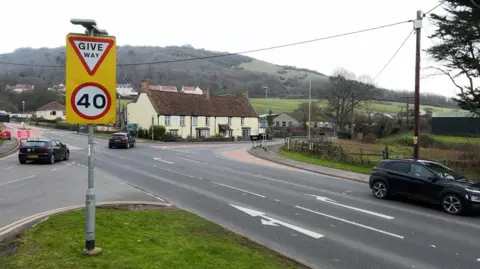 Big sign saying 40mph and Give Way. It's a road with four separate exits with cars moving through the junction. The Mendip Hills are in the background.