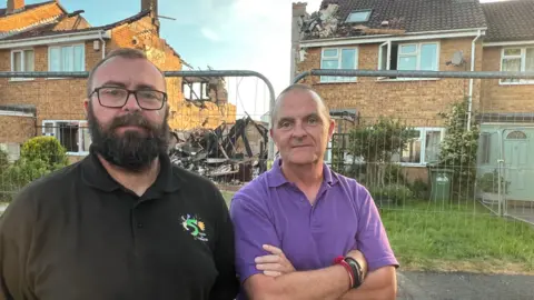 Two men standing in front of a fence, which is blocking access to a street of houses including one which was completely destroyed by a fire