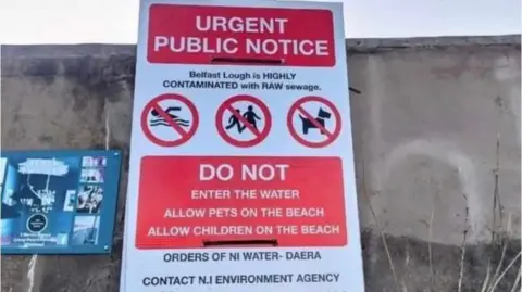 A red and white poster that say "urgent public notice", and describe Belfast Lough as "highly contaminated with raw sewage" and tell people not to go into the water or allow pets and children onto the beach.