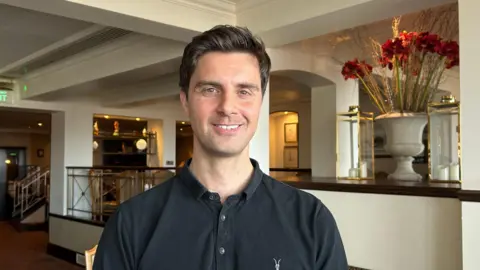 Matt Seymour, a man with short brown hair, is wearing a dark collared shirt. He is pictured in a hotel lobby. To the right of the image is a large vase of red flowers.