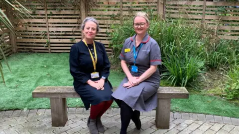 Joanne White on the right is wearing a nurse's uniform and glasses. Her blonde hair is tied in a bun. She is sitting on a bench in a garden next to Claudia Hepworth who is older with grey hair and wearing a dark skirt and top with dark red tights