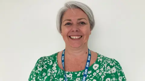 A woman is smiling into the camera. She has short grey hair and is wearing a green and white floral dress and an NHS lanyard is around her neck. She's standing with a white wall behind her.