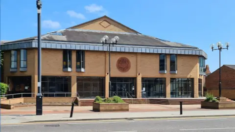 A photo of the modern brick building which has extensive black windows on the ground floor and a pitched roof. Steps surround the building and a CCTV pole is in the foreground of the picture.