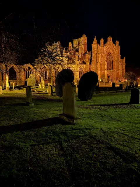 Ouraina Coleman An abbey with the building it up, and the surrounding graves in darkness