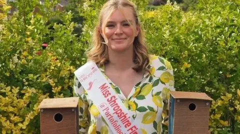 Nell Massin A woman with shoulder-length blonde hair wears a white dress with lemons on it. She is holding a bird house in each hand, and a sash that reads "Miss shropshire finalist 2025"