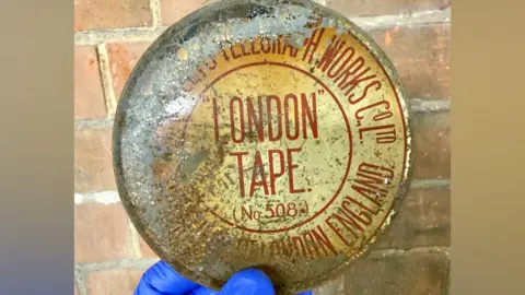 North Northamptonshire Council A metal round box, with writing in it, with rust marks. Some of the colour and writing has worn off. It is being held up by a brick wall and a person is holding it wearing blue gloves. 