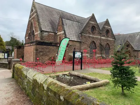 An old sandstone building with a number of peaks to its roof sits in gardens with a tree and a wall around it with a green sign for a cafe in front of the building