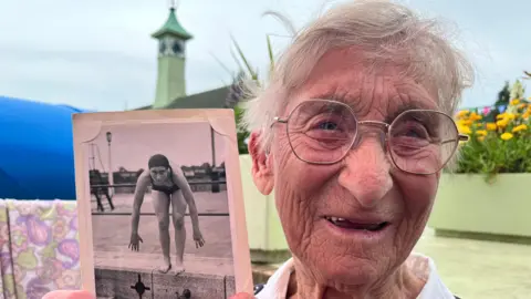 Pamela Cole is holding a picture next to her face, which is of her in the 1940s about to jump into water. Cole has grey hair and is wearing gold framed glasses and a white shirt. She is standing in front of plants.
