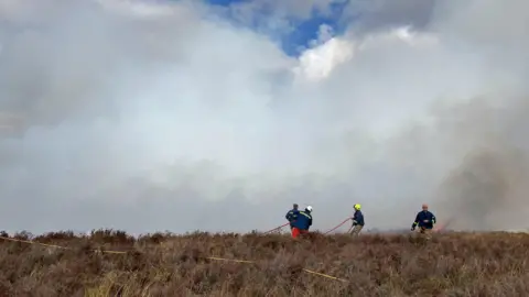 Staffordshire County Council A group of firefighters using hoses on a smoke-filled heathland during a wildfire.