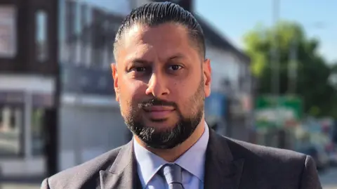 Shaz Saleem facing the camera. He has black hair and a black beard and moustache. He is wearing a grey suit with a blue shirt and tie.