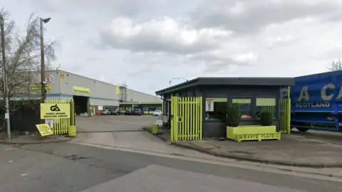 Street view of the site on an industrial estate with neon gates and a large grey metal-clad building in the background.