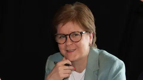 Nicola Sturgeon sitting down, wearing a pale green suit over a white top. She has her hand underneath her chin and is holding a pen.