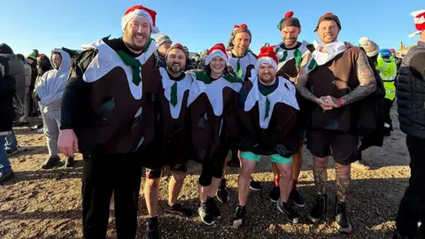 Everyone Active A smiling group of people dressed as Christmas puddings stand together on the beach before entering the sea