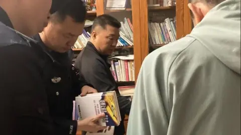 Sean Long / CSW Police officers thumb through books as Sun Cong dressed in black looks on
