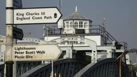 Natural England Signposts indicating the way to the King Charles III England Coast Path and other attractions at Sutton Bridge. In the background is the Cross Keys Bridge, a white and grey metal swing bridge with a Victorian engine house on top. 