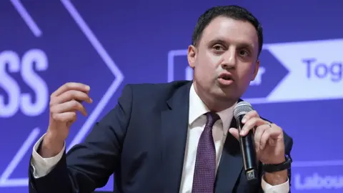PA Media Anas Sarwar wearing a dark suit over a white shirt with a purple tie. He is holding a microphone to his mouth. He is sitting in front of a purple background with white writing.