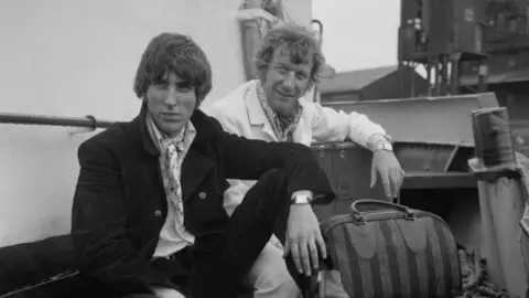 Getty Images A black and white photograph of disc jockeys Johnnie Walker (left) and Robbie Dale, of ship-based pirate radio station Radio Caroline South, at Felixstowe after the British government outlawed the station under the Marine Broadcasting Offences Act, 14 August 1967. They are both sitting with luggage and are looking directly at the camera.