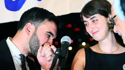 Mamdani is seen near a microphone on stage. He is wearing a suit with a tie and is kissing the hand of his wife, who is wearing a black dress with gold jewellery. 