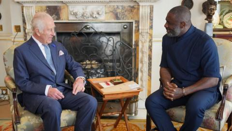 The King talking to Idris Elba in a room at St James Palace, they're sat next to each other on chairs and are chatting to each other
